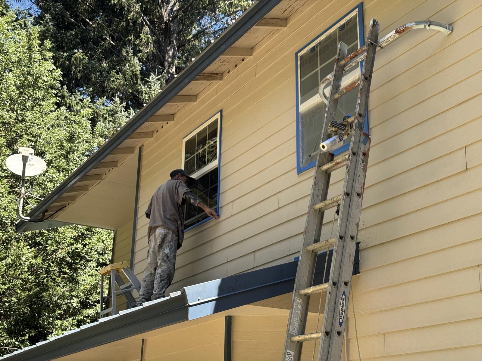 A painter cutting in the trim around an upstairs window by hand, standing above the porch roof with an extension ladder beside him.