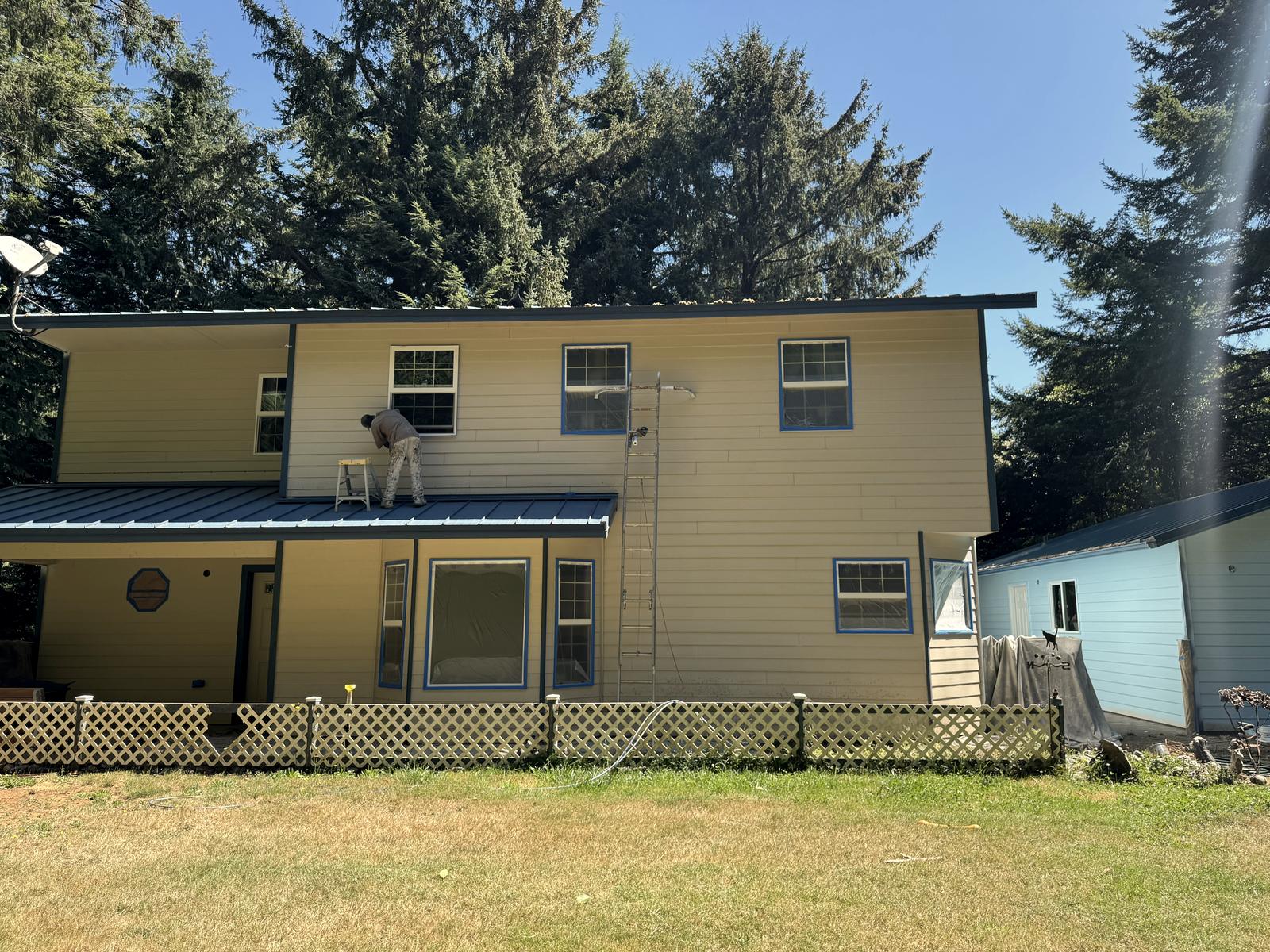 The back of the house part way through, a painter working above the porch roof with the windows masked off in blue.