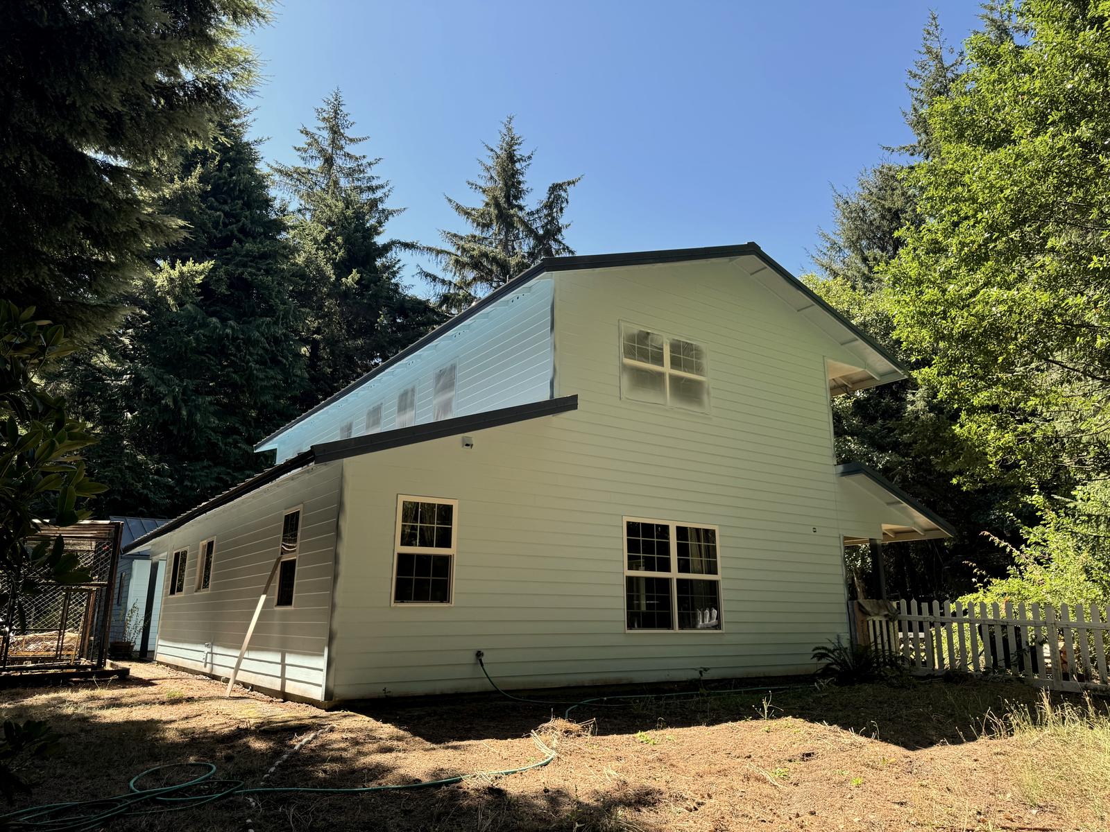 A finished side of the house in pale cream, a white picket fence running along the corner and firs standing behind it.