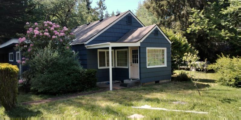 The finished cottage from the front, deep navy shingles with white trim and a rhododendron in flower beside it.