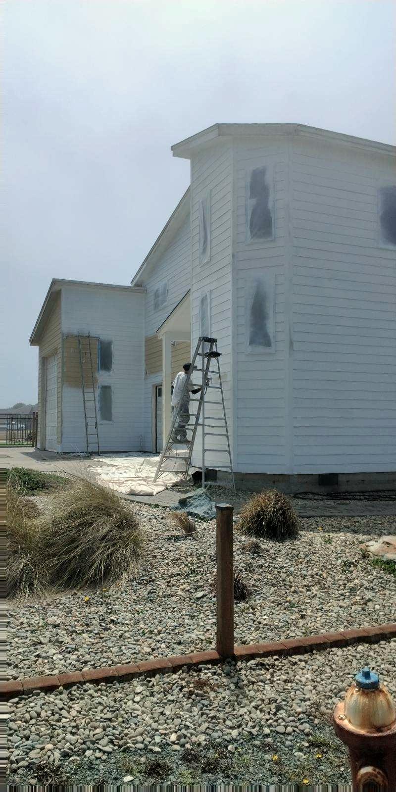 A two-story house primed in white on a foggy morning, a painter on a step ladder at the corner and the windows masked off.